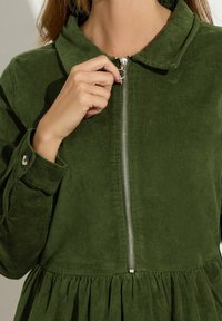 Groene corduroy jurk met een kraag en een rits aan de voorkant. Heeft een geplooide rok en mouwen met manchet en knoopdetails. Soepele textuur.