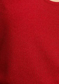 Roter gerippter Strickstoff mit subtiler Schimmer. Hat einen runden Halsausschnitt und ein strukturiertes Erscheinungsbild, das vertikale Linien betont.