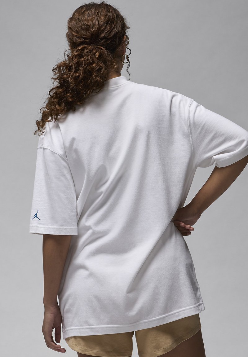 T-shirt bianca oversize realizzata in cotone con maniche corte. Presenta un piccolo logo blu sulla manica sinistra. Sfondo grigio.
