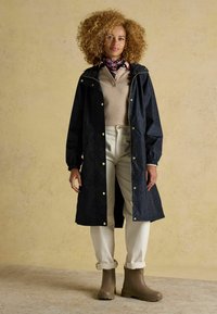 Impermeabile blu navy con bottoni a pressione dorati e zip, indossato sopra un maglione beige e pantaloni color crema. Stivaletti marroni impermeabili completano l'outfit.