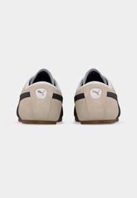 Ζεύγος μπεζ σουέντ αθλητικών παπουτσιών Puma με μαύρες ρίγες, εμφανιζόμενα από πίσω σε λευκό φόντο.