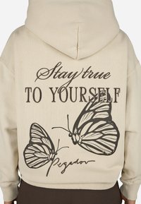 Κρεμ μπλουζάκι με κουκούλα που διαθέτει μεγάλης έκτασης εκτύπωση δύο πεταλούδων και το κείμενο "Μείνε πιστός στον εαυτό σου" σε μαύρο χρώμα.