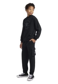 Μαύρη μπλούζα με κουκούλα και μπροστινή τσέπη, με λογότυπο αστεριού σε τονικές αποχρώσεις. Συνδυασμένη με μαύρα cargo παντελόνια και μαύρα αθλητικά παπούτσια με λευκές κορδόνες.