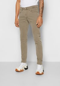 Beige slim-fit jeans med fem fickor, dragkedja och metallknapp. Bärs tillsammans med vita och svarta sneakers med texturerade gummisulor.
