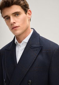 Marineblauer Wollmischmantel mit strukturierter Oberfläche, in doppelt geknöpftem Design und kariertem Muster in Braun. Auffällige spitze Revers.