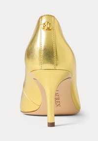 Gold high-heeled pump with a sleek design, metallic finish, pointed toe, and a small logo accent on the heel. Smooth texture.
