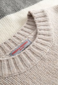 Lichtbeige gebreide trui met geribde kraag, voorzien van een grijze label met de tekst "FRIEDA & FREDDIES", gestructureerde stof en een zachte uitstraling.