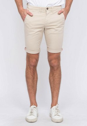 Homme portant un short beige retroussé, des baskets blanches et un t-shirt blanc rentré, les mains dans les poches.