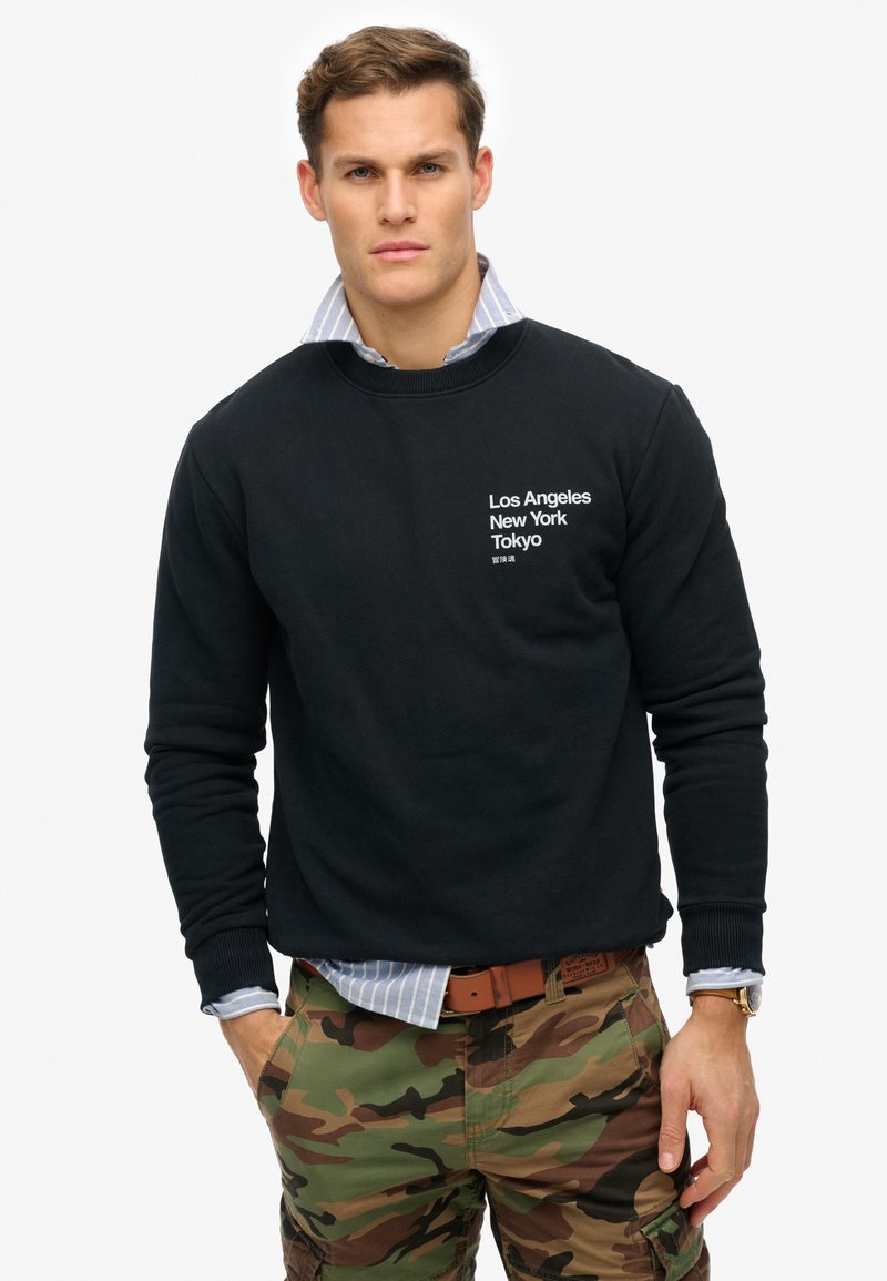Sudadera negra con cuello redondo, con texto en blanco: "Los Ángeles Nueva York Tokio." Combinada con pantalones cargo de camuflaje y una camisa de rayas por debajo.