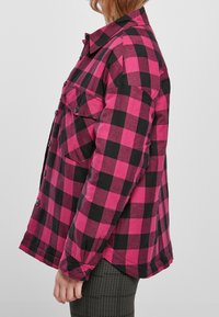 Camisa de franela a cuadros rosa y negra con cierre de botones y dos bolsillos frontales. Corte oversized con dobladillo recto y mangas relajadas.