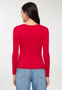Czerwona bluzka z długim rękawem, wykonana z gładkiej tkaniny, z okrągłym dekoltem i dopasowanym krojem. Połączona z jasnobłękitnymi dżinsami z wysokim stanem.