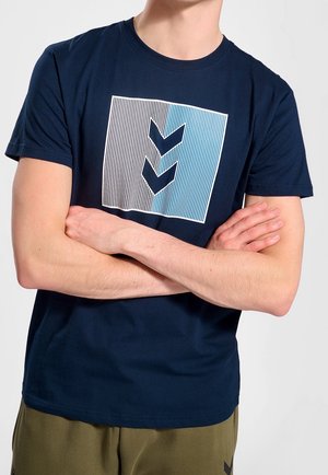 Personne portant un t-shirt bleu marine avec un carré rayé vertical bicolore et deux chevrons orientés vers le bas, les bras croisés sur la poitrine.