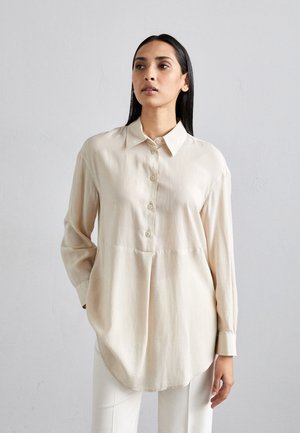Camicia beige con bottoni, dal tessuto morbido, colletto classico e maniche lunghe. Presenta un orlo curvo e un delicato dettaglio di piega frontale.