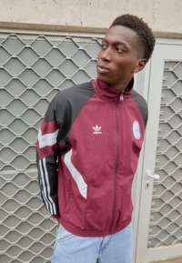 Casaco leve em marrom e preto com riscas brancas, gola alta e logótipo da Adidas; apresenta punhos elásticos e fecho de correr na frente.