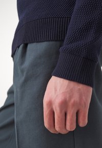 Stickad marinblå tröja med texturmönster och ribbade ärmslut, tillsammans med mörkgröna bomullsbyxor. En hand vilar synligt på byxorna.