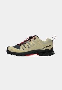 Zapatos de senderismo Salomon en color beige claro con acentos en negro y rojo. Presentan una parte superior texturizada, un sistema de ajuste con cordón y una suela resistente para una mejor tracción.