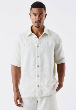 Camisa de manga corta en color crema claro, fabricada con un tejido texturizado, con un cierre de botones en la parte delantera, que presenta botones grises y un diseño de cuello clásico.