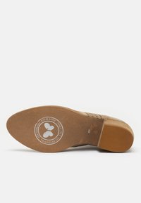 Semelle de chaussure en daim couleur fauve avec un design moulé, présentant un logo circulaire au centre. Forme ovale avec une prise texturée et des détails de taille et de marque en relief.