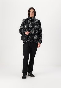 Svart og hvit paisley-mønstret fleecejakke, halv-zip krage, kombinert med svarte bukser og en liten svart skulderveske. Mørke sneakers.