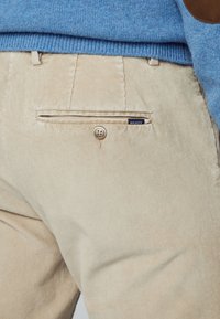 Beige kordflodbyxor med vertikala ribbor, med en knappad bakficka och en liten blå och vit etikett som står "HACKETT."