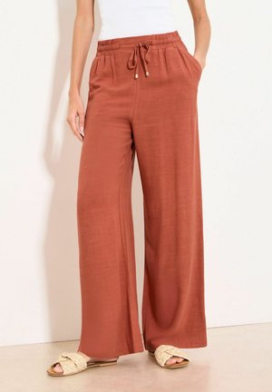 Rust-colored wide-leg pants with elastic waistband and drawstring, worn with white top and beige braided flat sandals.