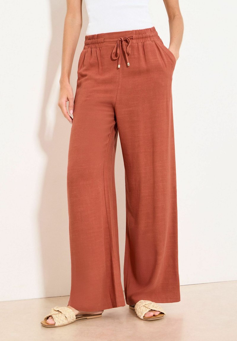 Pantalon large couleur rouille avec taille élastique et cordon de serrage, porté avec un haut blanc et des sandales plates tressées beiges.