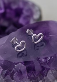 Silver hjärtformade stiftörhängen med en öppen design, som sitter ovanpå lila kristall. Slät yta med subtila reflektioner och transparens.
