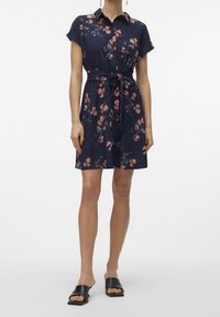 Kvinna iklädd en marinblå klänning med kort ärm och bälte, prydd med rosa blommönster, samt svarta sandaler med öppen tå, stående framför en vit bakgrund.