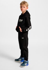 Sort hættetrøje med "hummel" logo og hvidt chevronmønster på ærmerne, kombineret med sorte joggers og blå sportssko.