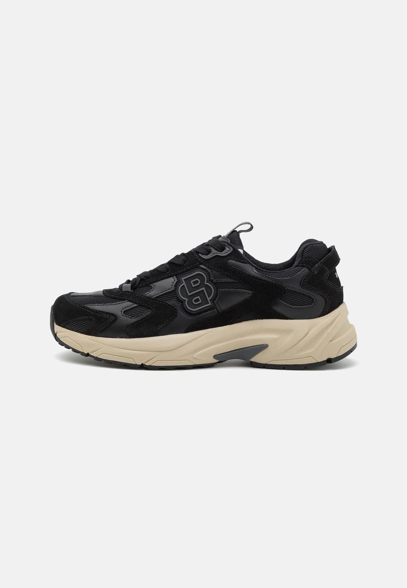 Chaussure de sport noire avec semelle épaisse beige, fermeture à lacets et logo stylisé sur le côté sur un fond clair uni.