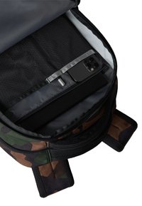 Wnętrze plecaka w czarno-brązowym kamuflażu; zawiera szare wykończenie, siatkową kieszeń, małą kieszeń na zamek oraz telefon spoczywający na wyściełanej części.