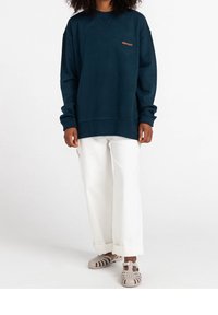 Felpa oversize blu navy con collo a giro e polsini a costine, dotata di un piccolo logo arancione. Abbinata a pantaloni bianchi a gamba larga e scarpe beige.