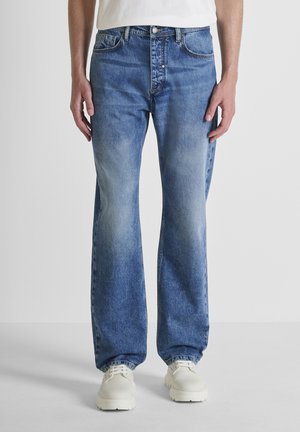 Man draagt lichtblauwe spijkerbroek met rechte pijpen en witte chunky schoenen met veters, staand tegen een effen grijze achtergrond.