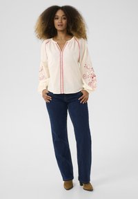 Crème blouse met rode borduursels en een geraffineerde halslijn, gecombineerd met donkerblauwe jeans. De blouse heeft lange, opgeblazen mouwen met decoratieve accenten.