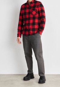Camisa de franela a cuadros rojos y negros con cierre de botones y bolsillo en el pecho, combinada con chinos grises y botas negras con cordones.