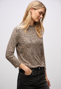 Beige Oberteil mit schwarz gesprenkeltem Muster, langen Ärmeln, runder Ausschnitt und weicher Textur; kombiniert mit hochgeschnittenen schwarzen Jeans.