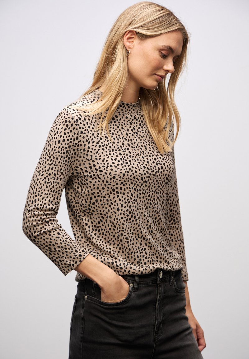 Beige Oberteil mit schwarz gesprenkeltem Muster, langen Ärmeln, runder Ausschnitt und weicher Textur; kombiniert mit hochgeschnittenen schwarzen Jeans.