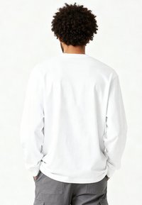 Man met krullend haar, gekleed in een eenvoudig wit shirt met lange mouwen en grijze cargobroek, staand met handen in de zakken, van achteren gezien.