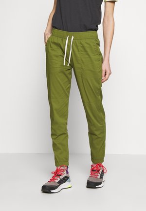 Persona que lleva pantalones verde oliva con cordón y bolsillos laterales, y zapatos deportivos multicolores con cordones rojo brillante, de pie frente a un fondo liso.