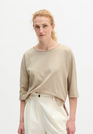 Kobieta o kręconych blond włosach nosi beżową koszulę z rękawem 3/4 wpuszczoną w kremowe spodnie z wysokim stanem, stoi na jednolitym białym tle.