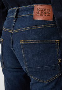 Tamno plave traperice s žutim šavovima i kožnom oznakom s natpisom "SCOTCH & SODA AMSTERDAM" na pojasu. Pravokutni stražnji džepovi.