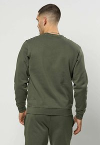 Mann med kort hår iført en olivengrønn genser med lange ermer og matchende bukser, stående med ryggen mot et ensfarget bakgrunn.