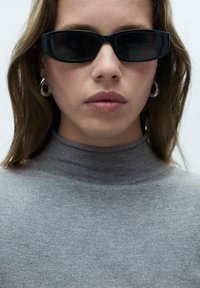 Čierne obdĺžnikové slnečné okuliare s matným povrchom, nosené cez šedý rolák, ktorý má hladkú textúru a priliehavý strih.