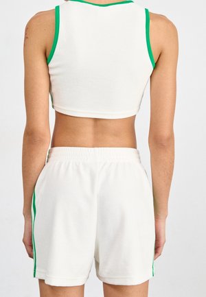 Persoon die een witte mouwloze cropped top en witte korte broek met groene bies draagt, gezien van achter tegen een effen achtergrond.