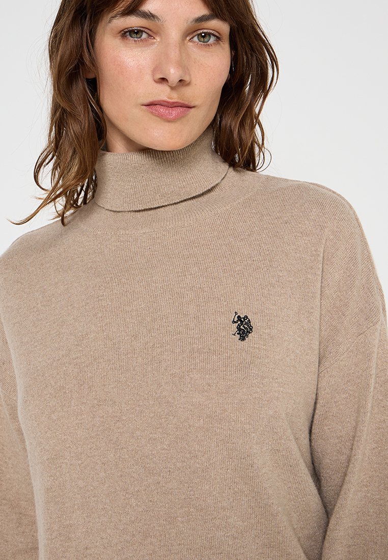 Maglione beige a collo alto realizzato in morbido tessuto a maglia, con un logo ricamato nero sul petto e colletto e polsini a costine.