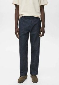 Donkerblauwe broek met een rechte pasvorm, gemaakt van een gestructureerde stof. De tailleband heeft riemlussen en het model draagt bruine schoenen.