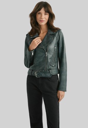 Kobieta o średniej długości brązowych włosach, ubrana w ciemnozieloną skórzaną kurtkę motocyklową i czarne spodnie, stojąca na tle jednolitego szarego tła.