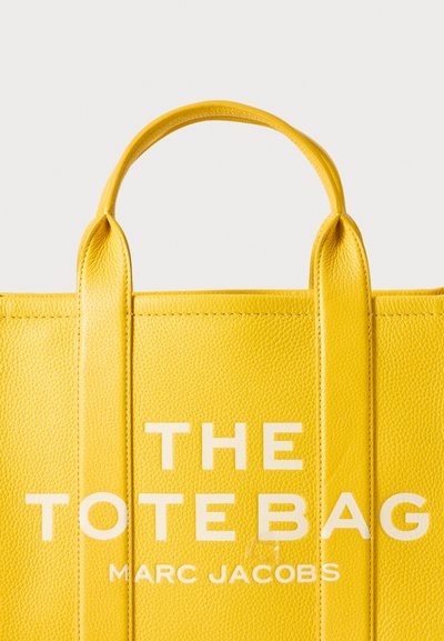 Yellow leather tote bag featuring two top handles, textured surface, and bold white text reading "THE TOTE BAG" and "MARC JACOBS".