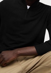 Schwarzer Zip-Pullover aus weichem Stoff, mit langen Ärmeln und gerippten Bündchen, kombiniert mit beigen Hosen und im entspannten Schnitt.