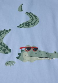 Sininen kangas esittelee sarjakuvamaisia alligaattoreita, jotka koristelevat punaiset aurinkolasit, teksturoidut vihreät suomut ja hymyllinen ilme.